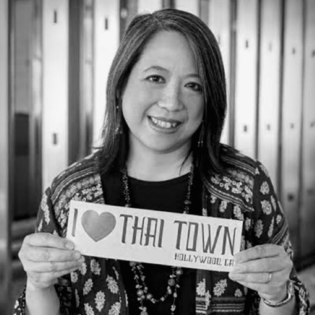 A woman smiles while holding a sign that reads I ♥ Thai Town Hollywood, CA. She is wearing a patterned jacket and necklace, and is standing in front of blurred lockers. The image is in black and white.