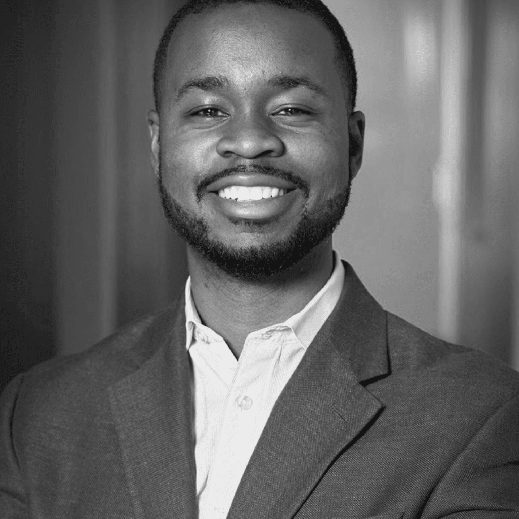 A man in a suit jacket and collared shirt smiles warmly at the camera in a black-and-white portrait, standing in front of a blurred indoor background.