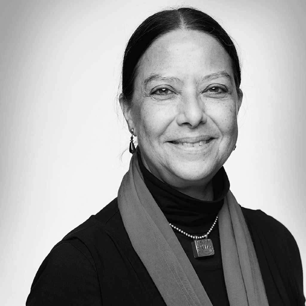 A woman with dark hair tied back smiles softly. She wears a dark top, a necklace, and a light scarf. The background is plain and the photo is in black and white.