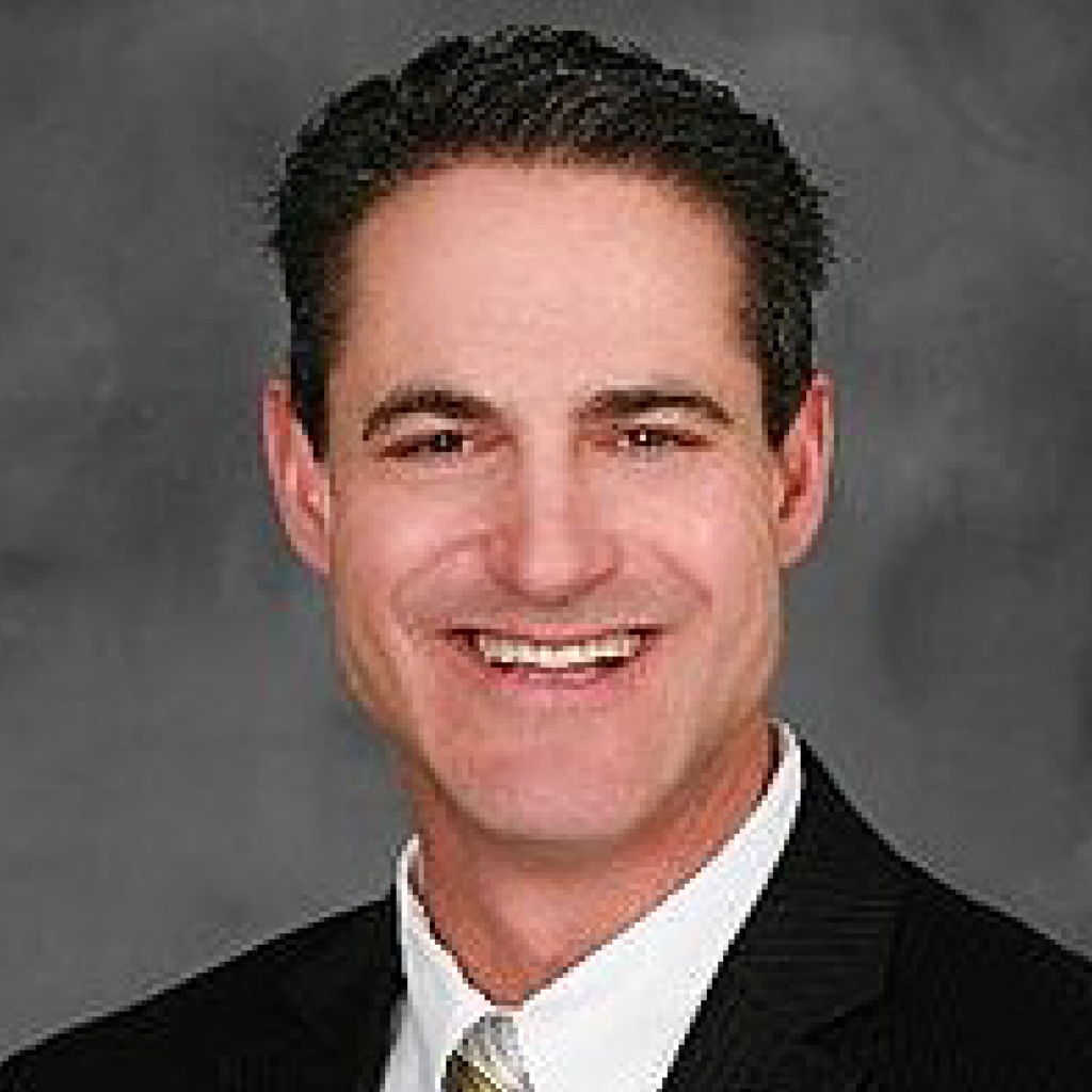 A man with dark, short hair wearing a black pinstripe suit, white dress shirt, and striped tie, smiling in front of a gray background.