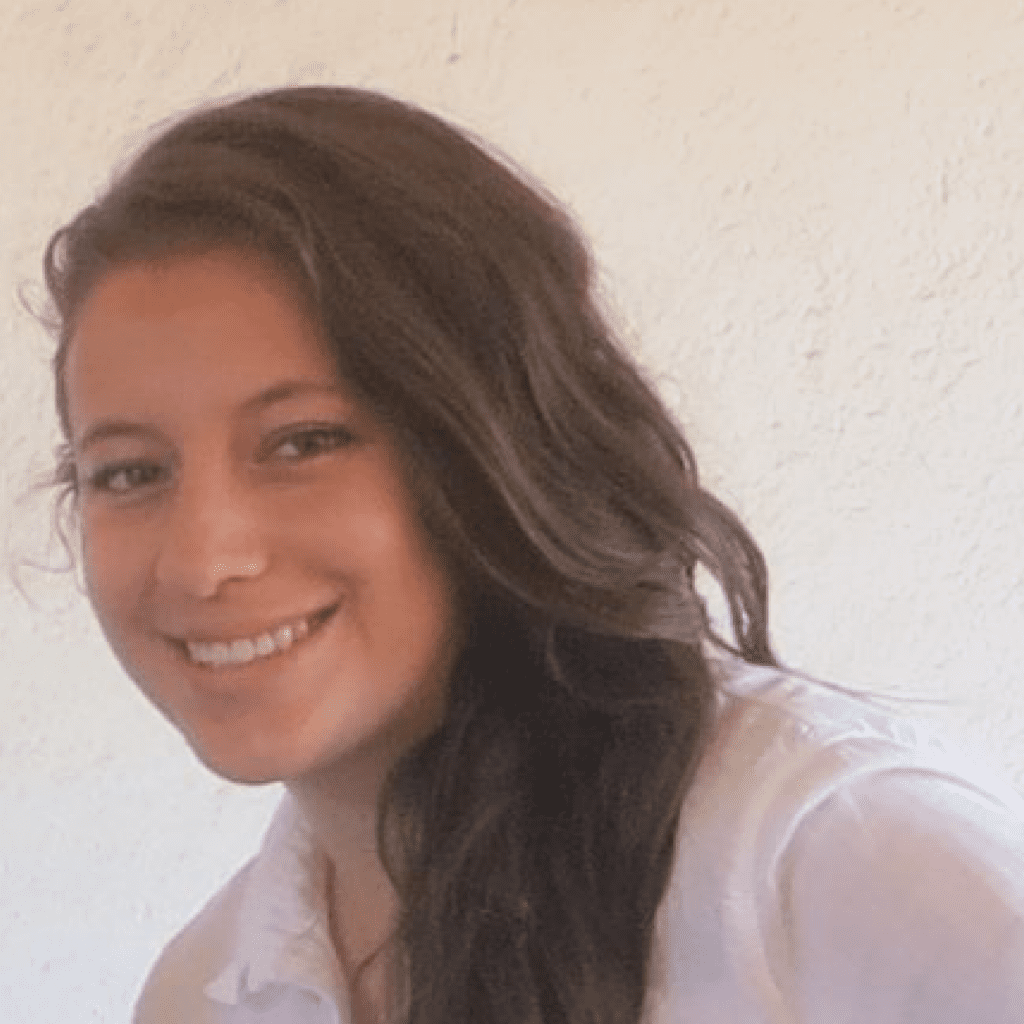 A young woman with long, wavy brown hair smiles while looking at the camera. She is wearing a white shirt and is positioned in front of a plain, light-colored wall.