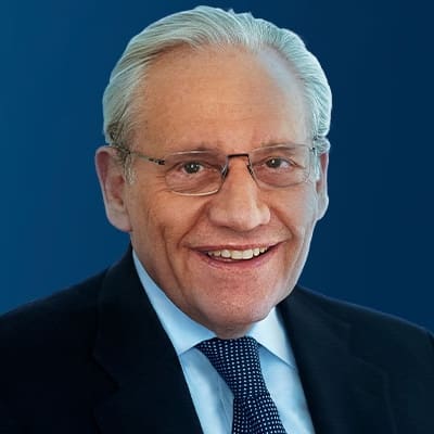An older man with gray hair and glasses smiles at the camera. He is wearing a dark suit, light blue shirt, and a navy polka dot tie. The background is a solid blue gradient.