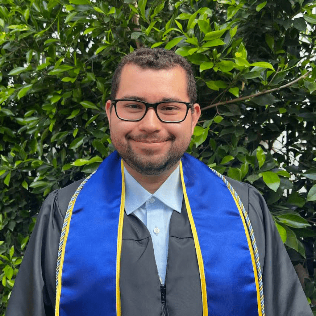 A person wearing glasses, a black graduation gown, and a blue stole with gold trim is smiling while standing in front of green leafy bushes.