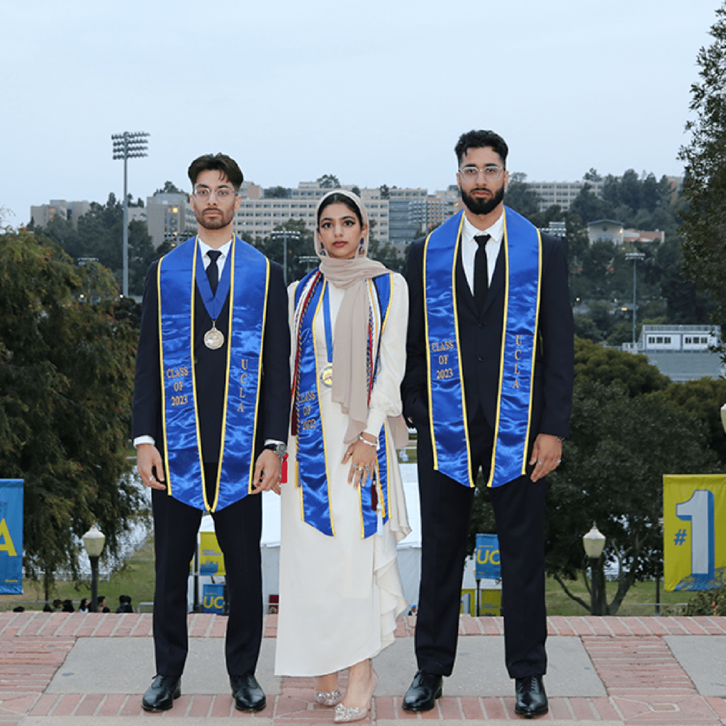 Three graduates stand side by side outdoors, wearing black suits, blue and gold graduation stoles, and medals. The central graduate wears a white dress and headscarf. Trees and buildings are visible in the background.