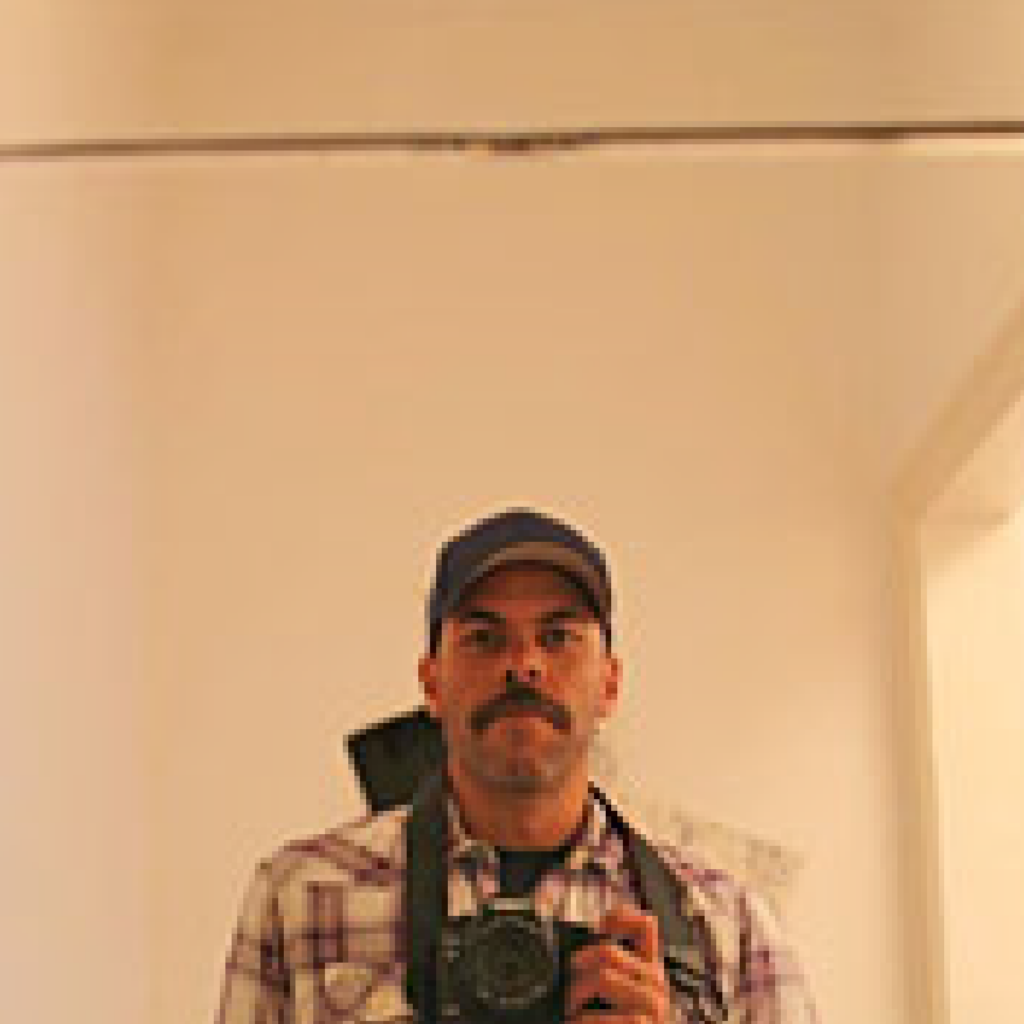 A person wearing a blue cap and plaid shirt takes a mirror selfie with a camera, standing indoors against a plain, light-colored wall.
