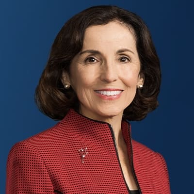 A woman with shoulder-length brown hair, wearing a red textured jacket with a black zipper and a small brooch, smiling in front of a solid blue background.