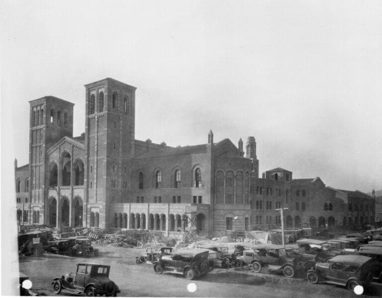 Historic black-and-white photo of a large Romanesque-style building under construction, with scaffolding and vintage cars parked in the foreground.