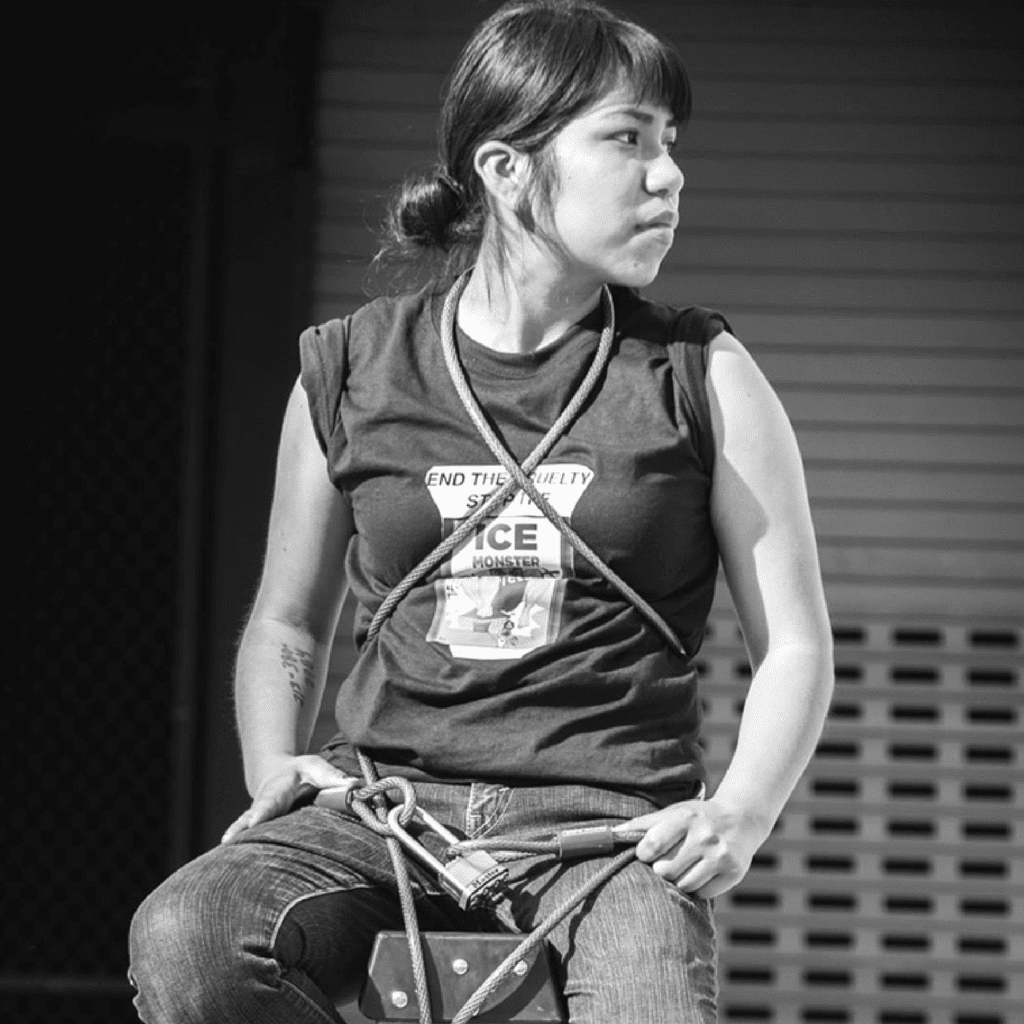 A person with dark hair in a ponytail sits, looking to the side. They wear a t-shirt with a visible protest message, ropes across their torso, and jeans. The photo is in black and white.