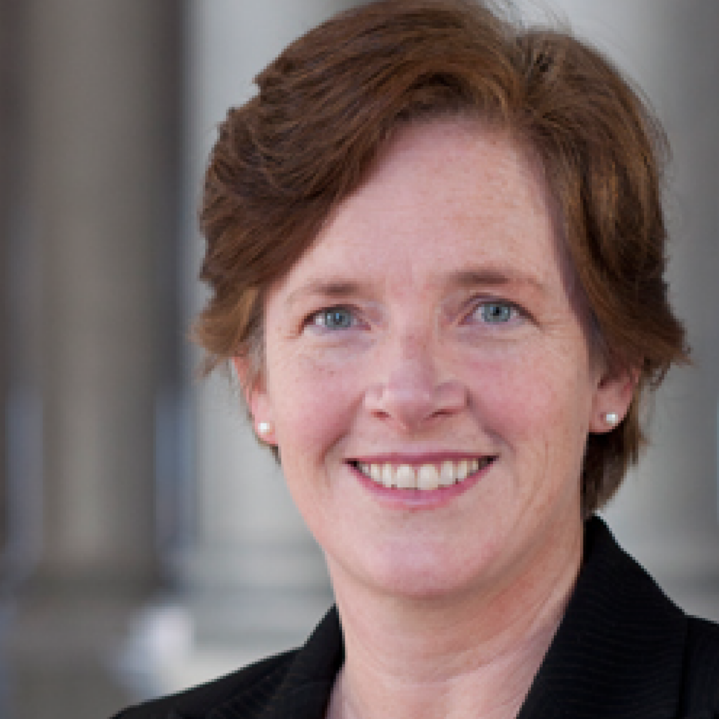 A woman with short, light brown hair wearing a black blazer and pearl earrings, smiling at the camera. The background is blurred with tall, light-colored columns.