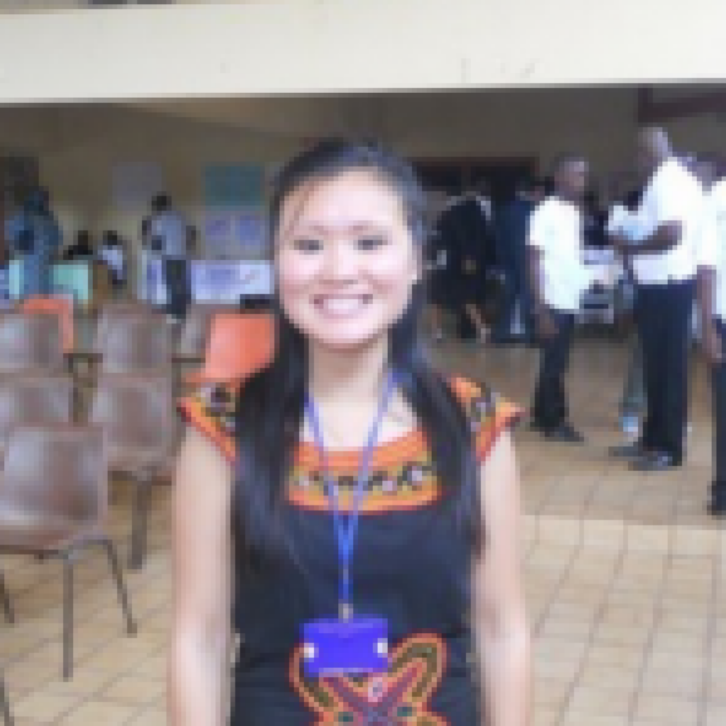A woman with long dark hair, wearing a black dress with colorful patterns and a blue lanyard, stands smiling indoors with chairs and people in the background.