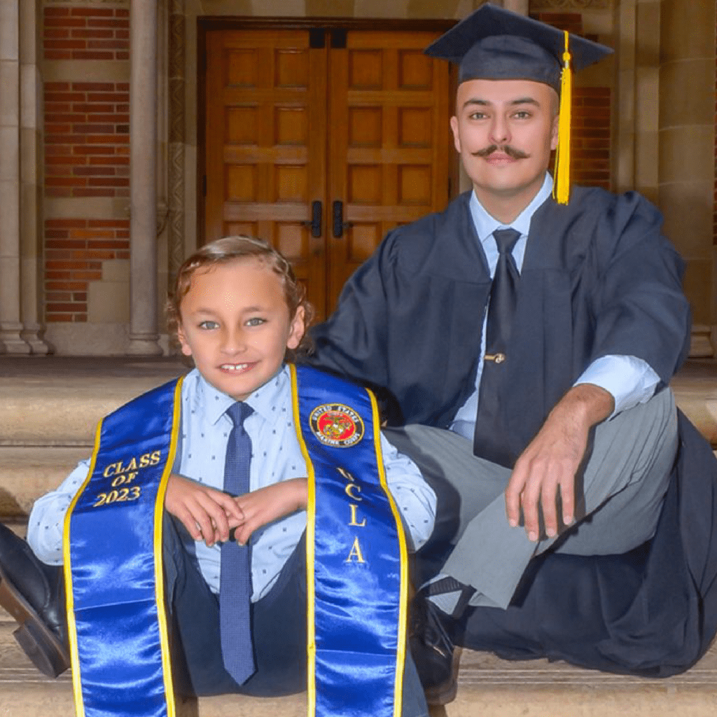 A graduate in a cap and gown sits on steps next to a smiling young boy wearing a shirt, tie, and UCLA graduation sash, both posing happily in front of a building.