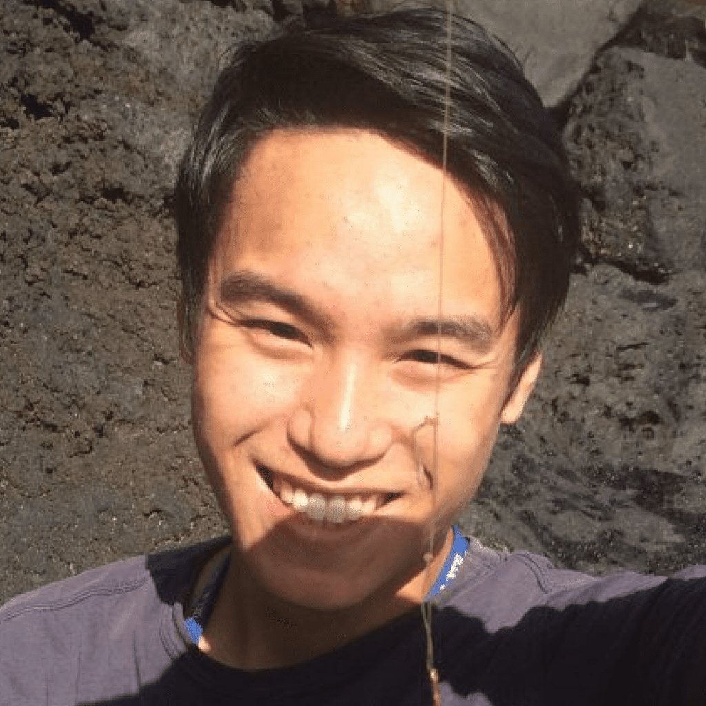 A person smiling outdoors with sunlight on their face and a thin plant stem crossing in front of them. Dark, rocky ground is visible in the background.