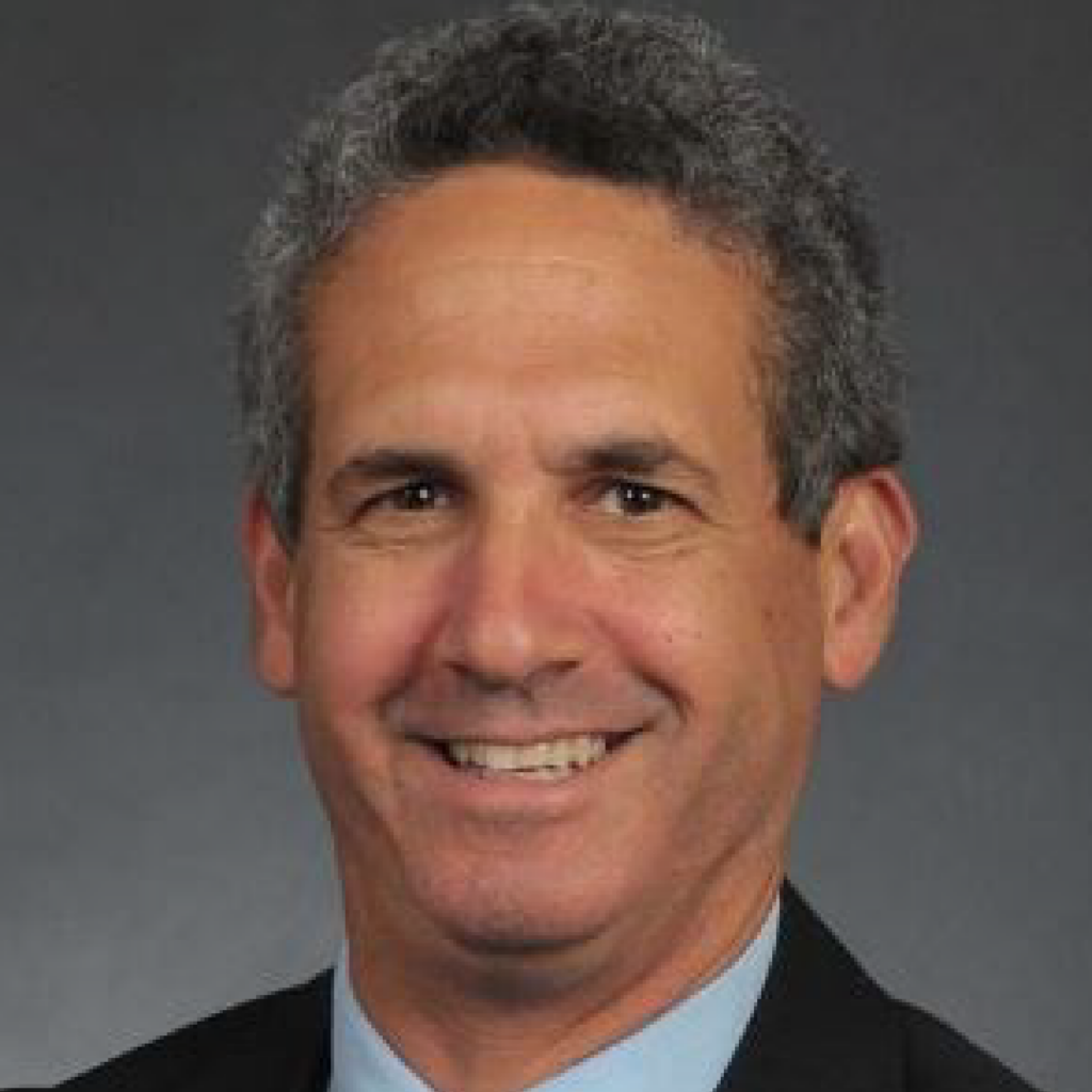 Portrait of a middle-aged man with short curly gray hair, wearing a dark suit jacket, light blue shirt, and smiling against a plain gray background.