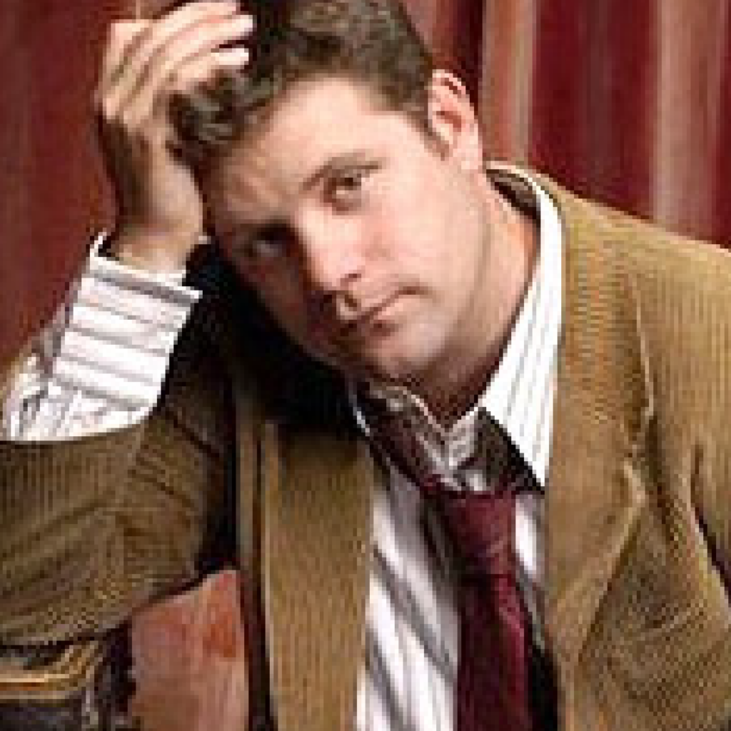 A man wearing a brown jacket, striped shirt, and loose tie rests his head on his hand, looking slightly pensive, with red curtains in the background.