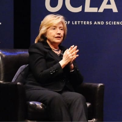 A woman in a dark suit sits on a black leather chair, clasping her hands, in front of a blue backdrop with the partial letters UCLA and the phrase Of Letters and Scienc visible.
