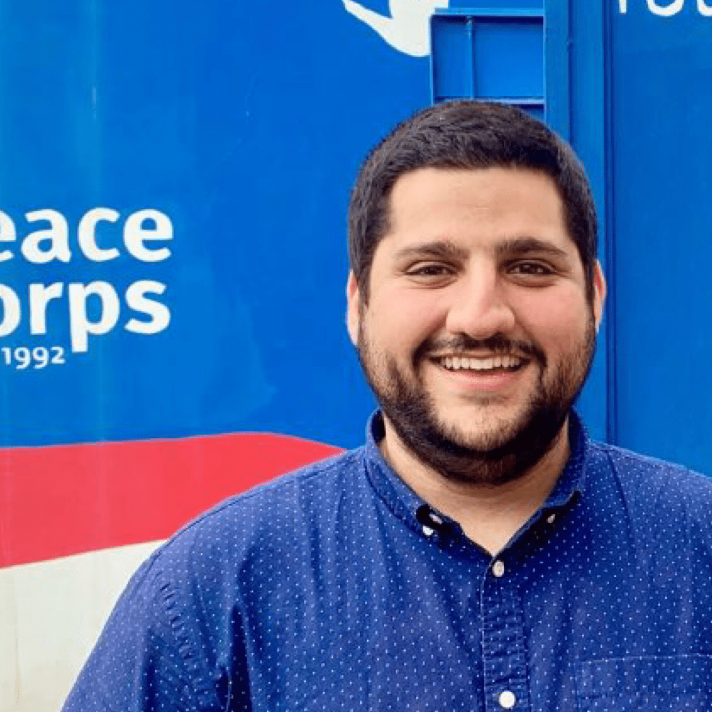 A smiling man with dark hair and a beard stands in front of a blue wall with the words Peace Corps partially visible in white text. He is wearing a blue patterned button-up shirt.