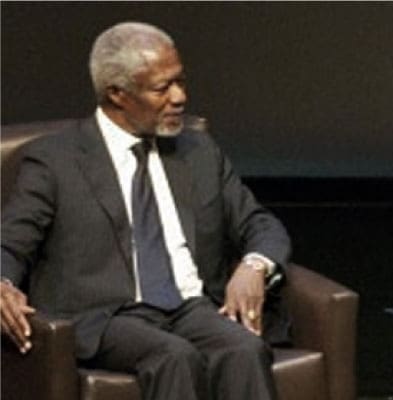 An older man with short gray hair and a beard sits in a brown armchair, wearing a dark suit, white shirt, and dark tie, looking to his left against a dark background.