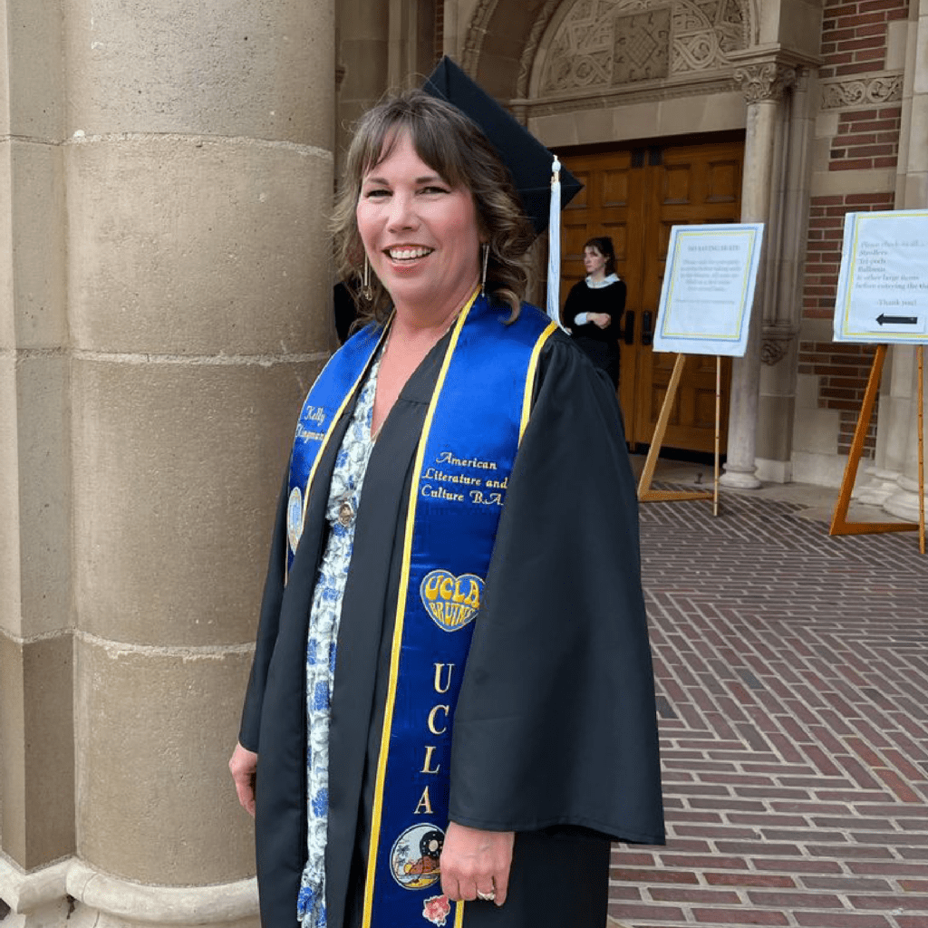 A woman in a graduation cap and gown stands smiling outside a building with large wooden doors. She wears a blue and gold UCLA stole and a patterned blouse under her gown.