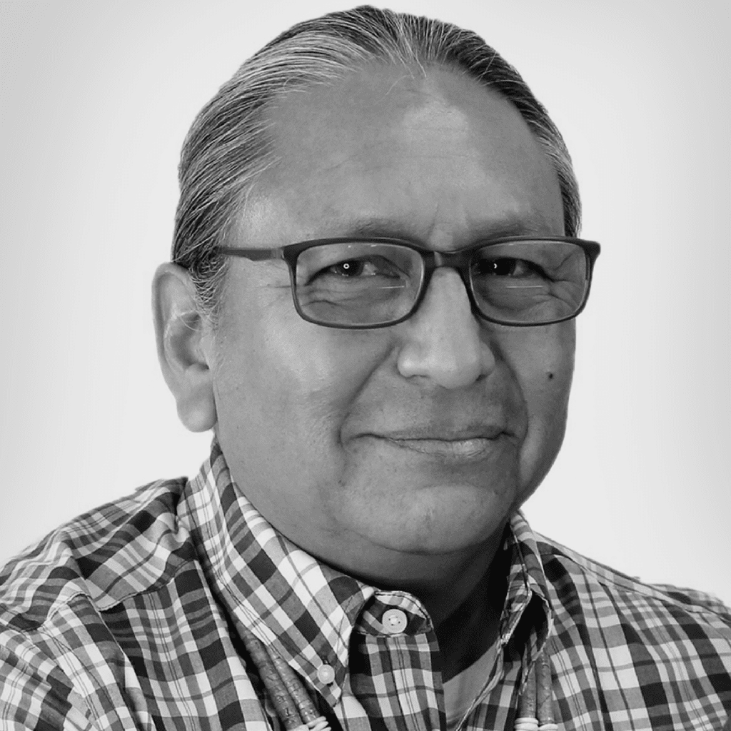 A man with gray hair and glasses smiles slightly, wearing a plaid shirt and a lanyard, posed against a plain light background.