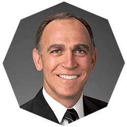 A middle-aged man with short brown hair, wearing a dark suit, white shirt, and striped tie, smiling at the camera against a gray background in an octagonal frame.