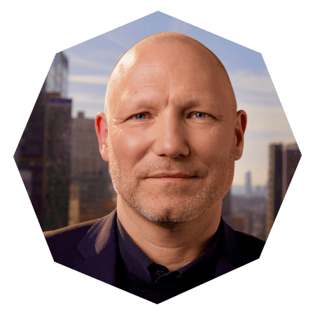 A middle-aged, bald man with light skin and blue eyes, wearing a dark jacket, stands in front of a cityscape with tall buildings under a clear sky. The photo is framed in a hexagonal shape.