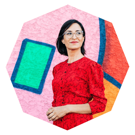 A woman with glasses wearing a red dress stands in front of a colorful, abstract mural with geometric shapes, looking slightly to the side and smiling.