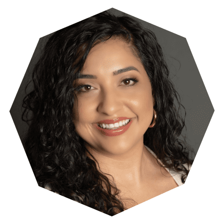 A woman with long, curly dark hair smiles at the camera. She is wearing hoop earrings and a light-colored top. The photo is cropped in an octagonal shape with a neutral gray background.