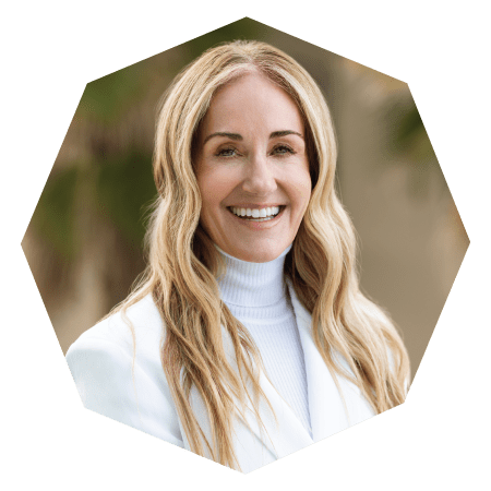A smiling woman with long, wavy blonde hair wearing a white turtleneck and white blazer, posed in front of a blurred outdoor background. The image is cropped in an octagonal shape.
