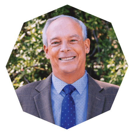A smiling older man with short gray hair, wearing a brown plaid suit jacket, blue dress shirt, and blue polka-dot tie, standing outdoors with trees in the background. Cropped in an octagonal shape.