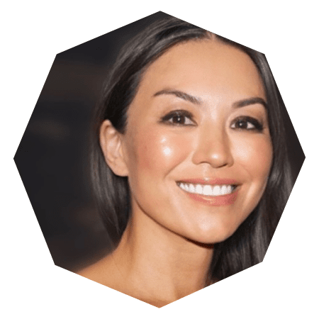 A woman with long dark hair and glowing skin smiles warmly. The image is cropped in an octagonal shape with a dark, blurred background.