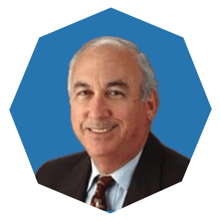 A middle-aged man with short gray hair, wearing a dark suit, light blue shirt, and patterned tie, smiling in front of a blue octagonal background.