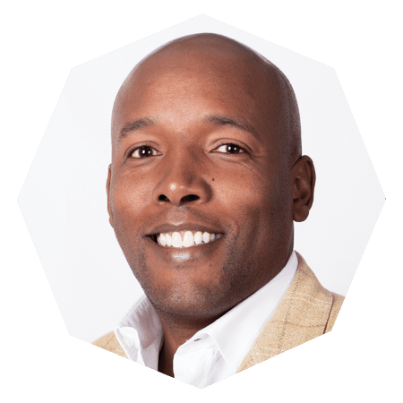 A smiling man with a shaved head wearing a beige jacket and white shirt, shown in a close-up portrait against a plain white background.
