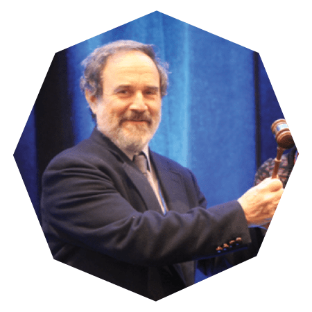 A man with gray hair and a beard, wearing a dark suit and tie, smiles while holding a wooden gavel against a blue curtain background.