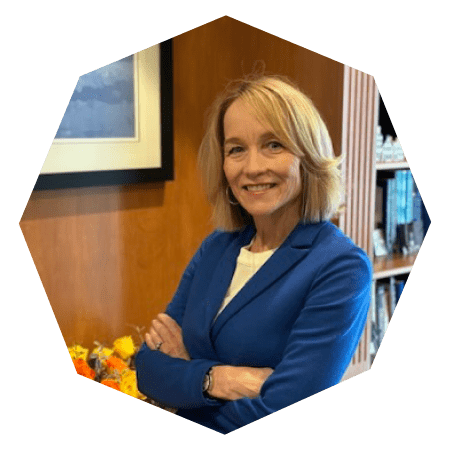 A woman with shoulder-length blonde hair, wearing a blue blazer and white top, stands smiling with arms crossed in an office setting with bookshelves and framed art in the background.