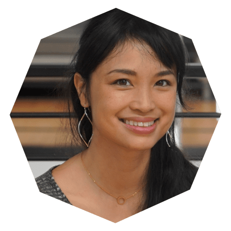 A woman with long dark hair smiles at the camera. She is wearing a pair of silver earrings and a delicate necklace. The photo is cropped in a hexagonal shape, and the background is softly blurred.