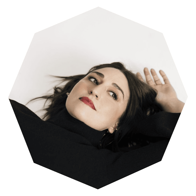 A woman with long brown hair and red lipstick lies on her back, head tilted, and arm raised above her head, looking off to the side. She wears a black turtleneck against a white background.