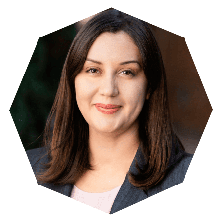 A woman with straight brown hair, wearing a dark blazer and light top, smiles softly at the camera. The photo is framed in a hexagonal shape with a neutral background.