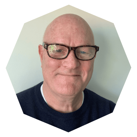 A smiling older man with glasses and a bald head, wearing a dark sweater, in front of a light-colored background. The image is cropped in a hexagonal shape.