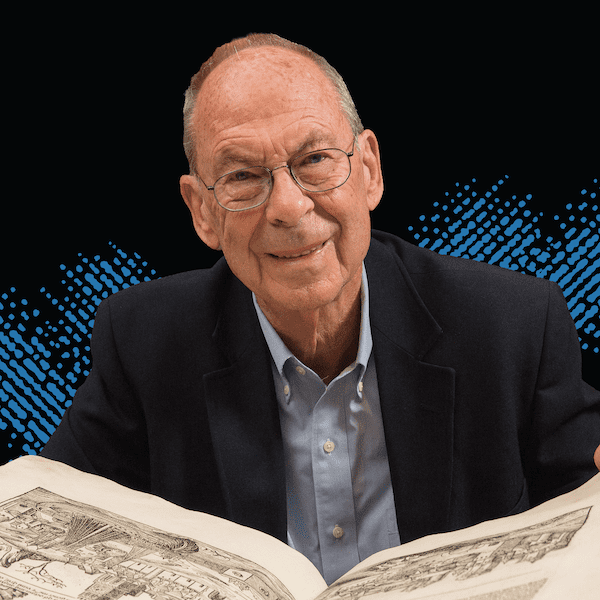 An older man wearing glasses and a dark blazer smiles while holding open a large illustrated book, with a blue dotted graphic pattern on a black background behind him.