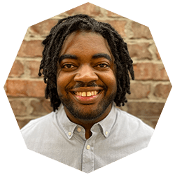 A man with medium-length, twisted hair and a beard smiles warmly at the camera, wearing a light-colored button-up shirt, standing in front of a brick wall.