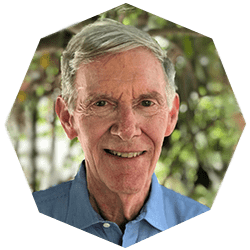 Smiling older man with short gray hair wearing a blue collared shirt, posed in front of greenery, seen in a hexagonal frame.