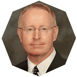 A middle-aged man with short light hair and glasses, wearing a dark suit, white shirt, and striped tie, is shown against a neutral background in a hexagonal-cropped portrait.