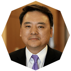 A man in a suit and purple tie is smiling slightly, facing the camera. He has short dark hair and is shown against a blurred brown background in a hexagonal frame.
