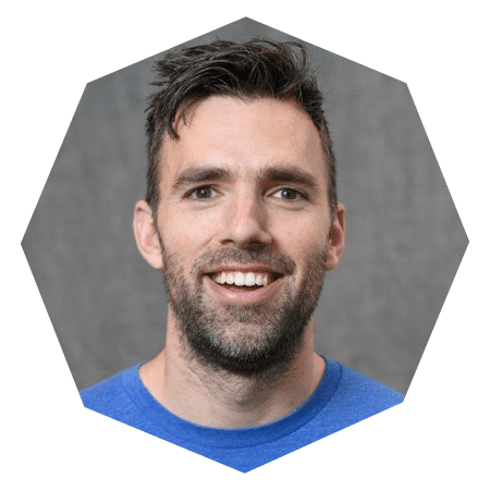 A man with short dark hair and a beard smiles at the camera. He is wearing a blue shirt and is photographed against a gray background, framed within a hexagonal border.
