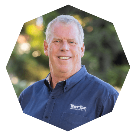 A smiling middle-aged man with short gray hair wearing a dark blue button-up shirt with the Yorke Engineering, LLC logo, standing outdoors with greenery in the blurred background.