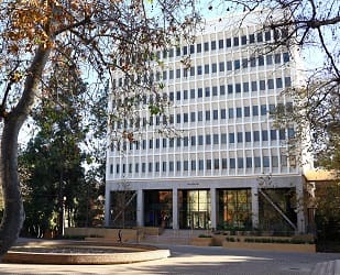 A tall, white, rectangular office building with many windows is partially framed by trees with autumn leaves. There is a brick plaza and people walking near the entrance of the building.
