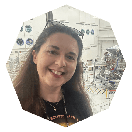 A woman with long brown hair and sunglasses on her head smiles at the camera in front of a large, bright room with machinery and equipment, possibly a space or science lab.