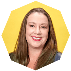 A woman with straight, long brown hair smiles in front of a bright yellow background. She is wearing a blue top and has light makeup with red lipstick. The image is framed in a geometric shape.