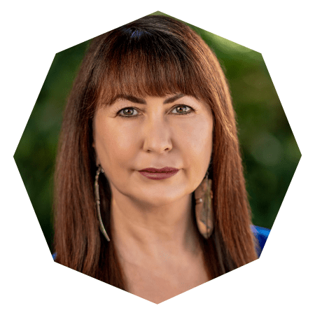 A woman with straight brown hair and bangs, wearing long earrings and a blue top, faces the camera with a neutral expression. The hexagonal photo has a blurred green background.