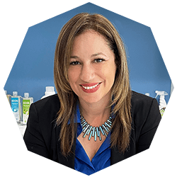 A woman with straight light brown hair, wearing a blue shirt, black blazer, and turquoise necklace, smiles at the camera. Behind her, bottles and spray containers are displayed on a shelf against a blue wall.